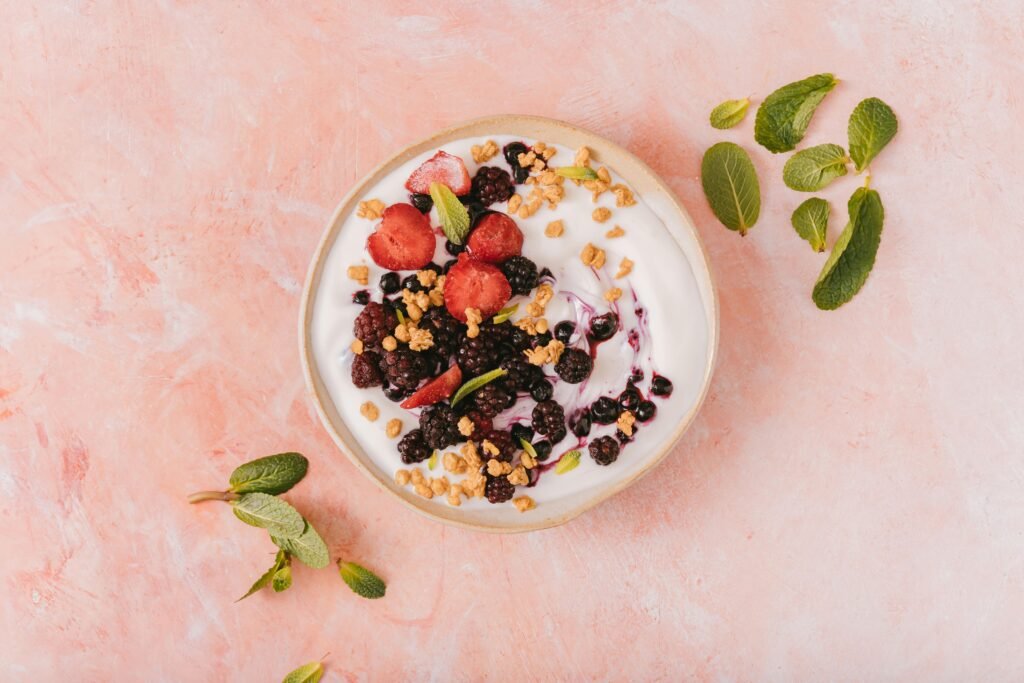 pexels photo 6823339 6823339 Top view of a vibrant yogurt bowl with mixed berries, granola, and mint leaves on a pink background.