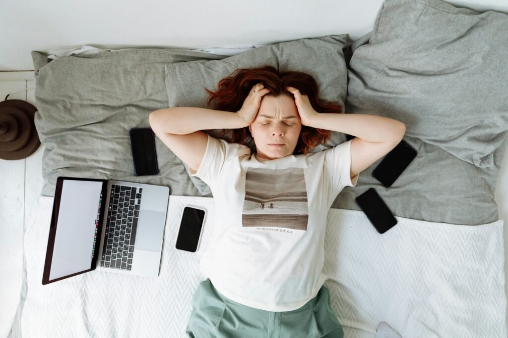 A distressed woman lying in bed surrounded by digital devices, showing signs of stress.
